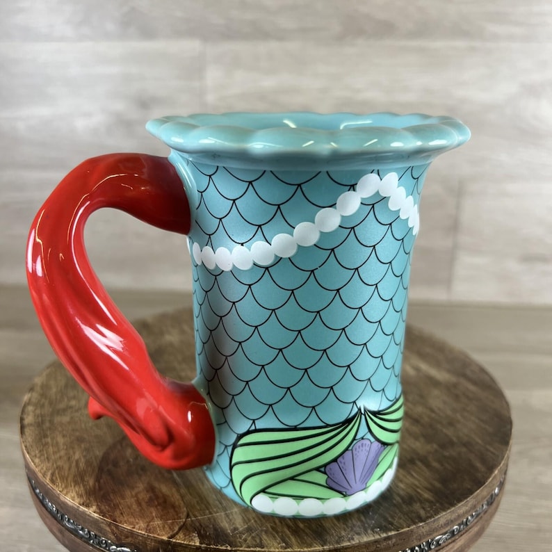 May include: A ceramic mug with a mermaid theme. The mug features a light blue body with a scale pattern, a red handle, and a white pearl necklace design. The base of the mug has a green and purple seashell design.