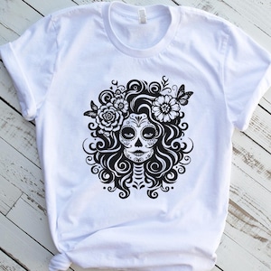 Puede incluir: Camiseta blanca con un dise&ntilde;o gr&aacute;fico en blanco y negro de una calavera de az&uacute;car con detalles florales y mariposas. El dise&ntilde;o presenta patrones detallados y un peinado decorativo.