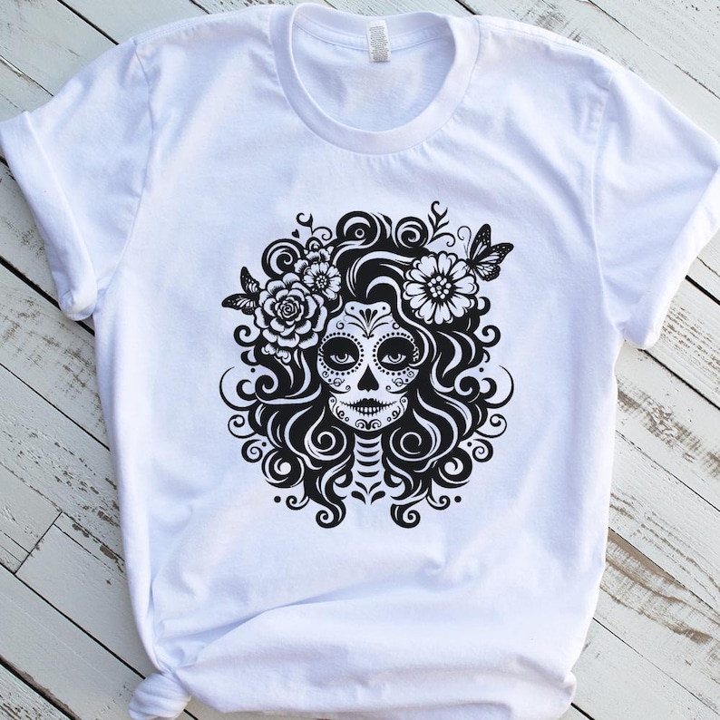 Puede incluir: Camiseta blanca con un dise&ntilde;o gr&aacute;fico en blanco y negro de una calavera de az&uacute;car con detalles florales y mariposas. El dise&ntilde;o presenta patrones detallados y un peinado decorativo.