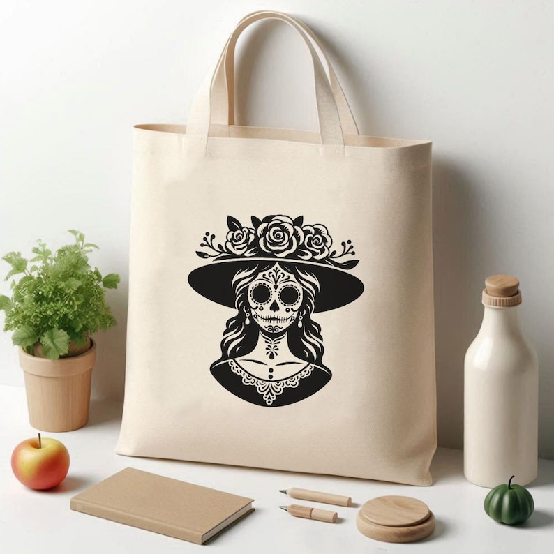 Puede incluir: Bolso tote beige con un gr&aacute;fico en blanco y negro de una mujer con maquillaje de calavera de az&uacute;car, que lleva un sombrero adornado con rosas. El dise&ntilde;o est&aacute; centrado en el bolso, con un asa sencilla. El bolso es de tela resistente.