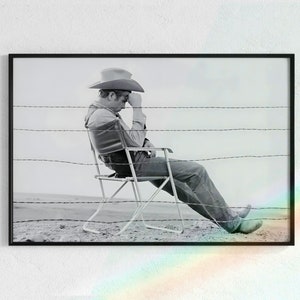 May include: A black and white photograph of a man wearing a cowboy hat sitting in a folding chair in front of a barbed wire fence.