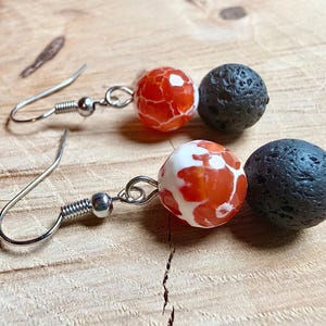 Op de afbeelding: Hangende oorbellen met een rood-witte patroonkraal en een zwarte, getextureerde kraal. De oorbellen hebben zilverkleurige oorhaken. De oorbellen worden gepresenteerd op een houten oppervlak.