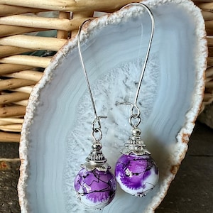 May include: A pair of dangle earrings with silver hooks and purple and white glass beads. The beads have a swirl design and are accented with silver.