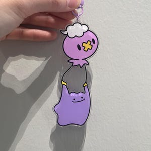 May include: A purple and white cartoon keychain featuring two characters. One character is a purple blob with a yellow cross and a cloud on its head. The other is a purple blob with a smiling face. The keychain has a purple clasp.