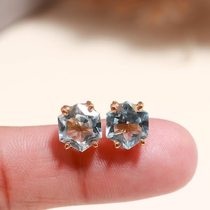 May include: A pair of hexagonal-cut, light blue gemstone stud earrings with gold-colored settings. The earrings are displayed on a finger, showcasing their delicate design and sparkling facets. The gemstones have a subtle, translucent quality.