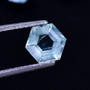 Puede incluir: Una piedra preciosa azul claro con un corte hexagonal, sostenida por unas pinzas sobre un fondo negro.