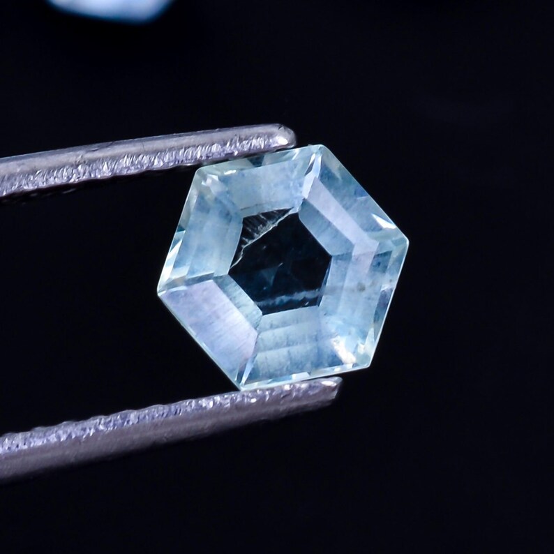 Puede incluir: Una piedra preciosa azul claro con un corte hexagonal, sostenida por unas pinzas sobre un fondo negro.