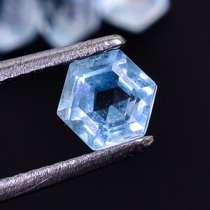 Puede incluir: Una piedra preciosa azul claro con un corte hexagonal facetado. La piedra preciosa se sujeta con unas pinzas sobre un fondo negro.