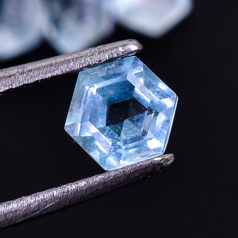 Puede incluir: Una piedra preciosa azul claro con un corte hexagonal facetado. La piedra preciosa se sujeta con unas pinzas sobre un fondo negro.