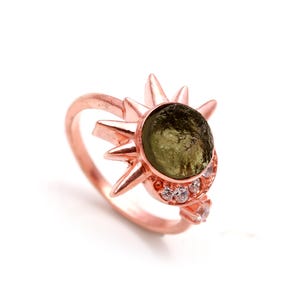 Könnte beinhalten: Ein Ring aus Roségold mit einem grünen Edelstein, der in einem Sonnenstrahl-Design gefasst ist. Der Ring ist mit kleinen, klaren Steinen verziert.
