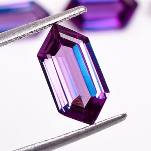 May include: A purple gemstone with a rectangular shape and a faceted cut. The gemstone is held by a pair of tweezers.