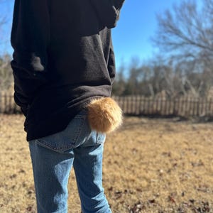 May include: A person wearing a black hoodie and blue jeans with a brown furry tail attached to the back of their jeans.