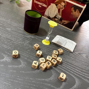 May include: A vintage Scrabble Crossword Cubes game set. The set includes a red box with a couple on the cover, letter cubes, a timer, and a score sheet. The letter cubes are scattered on a gray surface.