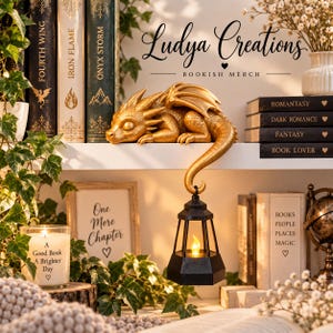 Può includere: Una statuetta di drago dorato con gli occhi chiusi che riposa su uno scaffale, con la sua lunga coda curva verso il basso che sostiene una lanterna esagonale nera contenente una luce calda. La scena include libri come Fourth Wing e Iron Flame.