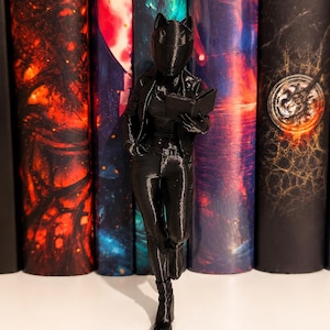 Peut inclure: Une figurine noire imprimée en 3D représentant un personnage portant un casque à oreilles de chat, une veste et un pantalon, debout tout en tenant un livre ouvert. La figurine a une finition lisse et monochrome, dans une pose décontractée.