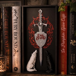Op de afbeelding: Een zwarte rechthoekige schaduwdoos met een zilveren zwaard en een schild met de tekst Dire Bound. Achter het zwaard bevindt zich een rood filigraanontwerp. Een witte wolf en een zwarte wolf zitten aan de voet van het zwaard.