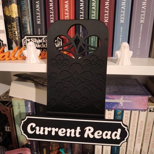 Peut inclure: Un support de livre en métal noir présentant un motif découpé complexe sur le dessus et une plaque rectangulaire à la base avec le texte blanc Current Read. Le support a une finition mate et un cadre droit pour exposer un livre.