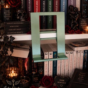 Könnte beinhalten: Ein salbeigrüner Buchständer aus Metall mit einem rechteckigen Rahmen und einer horizontalen Ablage für Bücher. Der Ständer hat eine glatte, matte Oberfläche und eine stabile Konstruktion, um offene Bücher aufrecht zu präsentieren.