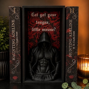 Könnte beinhalten: Ein schwarzes Buchregal-Display mit einer zentralen schwarzen Skulptur eines Oberkörpers mit Kapuze vor einem Hintergrund aus roten Rosen mit dem Text Cat got your tongue, little mouse? flankiert von zwei Hunting Adeline Büchern von H. D. Carlton.