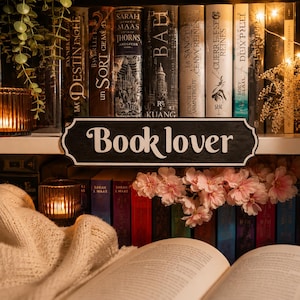 Peut inclure: Une enseigne en bois noir avec l'inscription blanche "Book lover" placée sur une étagère devant une rangée de livres. La scène comprend un tissu en maille crème, une petite bougie allumée dans un photophore et des fleurs roses.