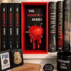Peut inclure: Un présentoir avec trois livres noirs intitulés The Mindf*ck Series de S.T. Abby, un cadre rouge contenant une sculpture de pomme rouge percée de clous, et divers objets dont un badge du FBI, un dossier confidentiel et une tasse noire avec texte.