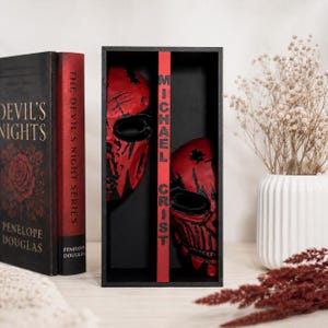 Peut inclure: Une boîte de présentation en bois noir contenant deux masques rouges et noirs avec des ouvertures pour les yeux. Le séparateur central porte le texte vertical MICHAEL CRIST en lettres noires. À gauche se trouve un livre noir intitulé Devil's Nights.