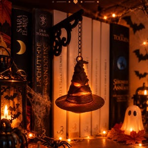 Peut inclure: Un chapeau pointu marron à larges bords avec des traits de visage, éclairé de l'intérieur, suspendu à une applique murale en métal noir par une chaîne. Le texte "Ludya Creations" et "BOOKISH MERCH" apparaît en haut de l'image.