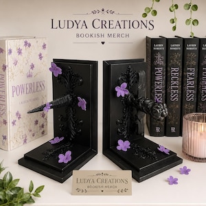 Peut inclure: Une paire de serre-livres noirs en forme de L avec des volutes ornées et des éléments centraux saillants, décorés de petites fleurs violettes. Un panneau rectangulaire devant indique LUDYA CREATIONS BOOKISH MERCH, assorti au logo au-dessus.