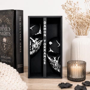 Peut inclure: Une boîte de présentation rectangulaire noire contenant deux moitiés de masque de crâne avec des détails blancs vieillis. Le séparateur central affiche le texte blanc vertical DAMON TORRANCE. Les moitiés sont dans des compartiments.