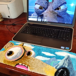 Op de afbeelding: Een zwarte laptop met een blauw en bruin kleed met een cartoonfiguur met grote ogen. Het laptopscherm toont een stilstaand beeld uit de animatiefilm Ice Age 2: The Meltdown. De tekst op het scherm luidt "Болезнь в очках - "Ледниковый период 2: Глобальное потепление" отрывок из фильма".