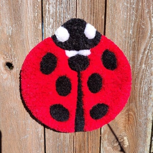 Può includere: Una coccinella rossa con macchie nere e dettagli bianchi è fatta di filato ed è attaccata a una superficie di legno.