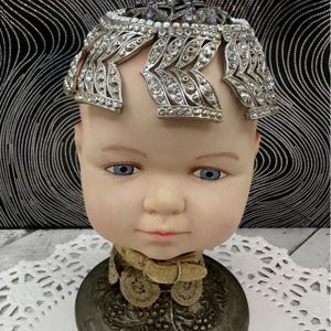 May include: A doll head with blue eyes and a light skin tone, wearing a silver-toned metal headpiece adorned with clear rhinestones in a leaf-like pattern. The doll has a tan-coloured lace collar with a small buckle around its neck.