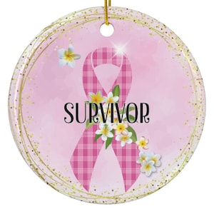 May include: Circular pink ornament featuring a plaid pink ribbon and the word "SURVIVOR" in black. The ribbon is adorned with white and yellow flowers. It has a gold hanging string.