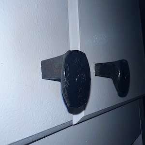 May include: Two black metal door handles mounted on a white wall. The handles are shaped like a hammer head with a rounded end.