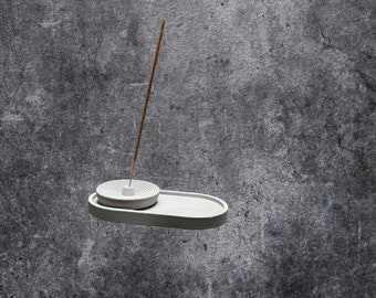 May include: A white ceramic incense holder with a round, raised center for holding incense sticks. The holder is on a gray textured background.