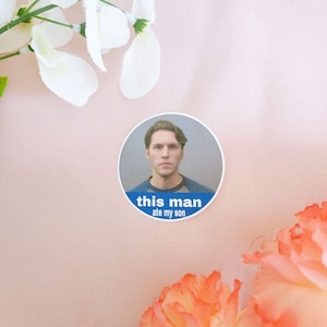 May include: A round sticker featuring a man's face inside a white circle. Below the image is a blue banner with the text "this man ate my son" in white. The sticker is on a pink background with white flowers.