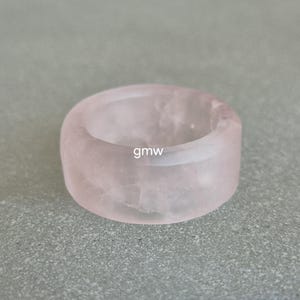 May include: A chunky, light pink rose quartz ring. The ring is a solid band with a smooth, polished surface. The text "gmw" is visible on the ring. This ring is a unique piece of jewelry.