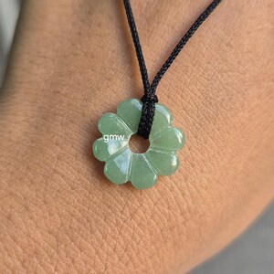 Puede incluir: Un collar con un colgante en forma de flor verde claro. El colgante tiene un agujero central y está ensartado en un cordón negro. El colgante está hecho de una piedra translúcida y pulida. Las letras "gmw" son visibles.