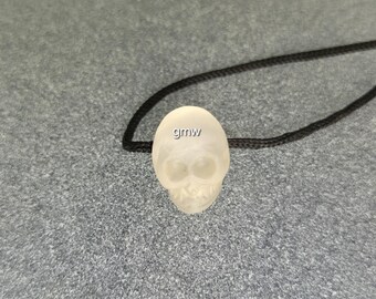 May include: A translucent skull pendant with a black cord. The skull has visible eye sockets and teeth. The letters "gmw" are visible on the top of the skull. The pendant is likely made of glass or a similar material.