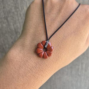 May include: Red flower pendant necklace with a black cord. The pendant is a carved flower shape with a hole in the centre. The pendant has the letters "gmw" on a small silver tag. The necklace is displayed on a hand.