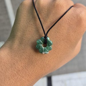 Puede incluir: Un collar con un colgante verde en forma de flor y un cordón negro. El colgante tiene un agujero en el centro y las letras "gmw" son visibles. El collar se sostiene en una mano.
