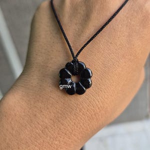 May include: Black flower-shaped pendant necklace with a black cord. The pendant has a circular design with a hole in the centre and the letters "gmw" on the side. The necklace is displayed on a person's hand.
