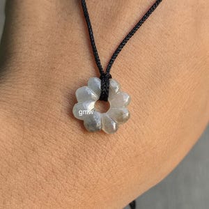 May include: A grey, flower-shaped pendant necklace with a black cord. The pendant has a carved floral design and the letters "gmw" are visible on the pendant. The necklace is displayed against a skin-toned background.