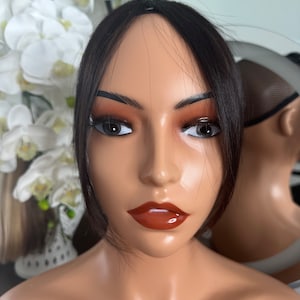 May include: A black hairpiece with bangs, designed to add volume and length to the front of the hair. The hairpiece is shown on a mannequin head with a neutral makeup look.