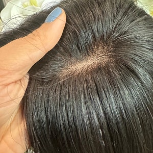 Puede incluir: Primer plano de una peluca de cabello humano marrón oscuro con un frente de encaje. La peluca está siendo sostenida por una mano con esmalte de uñas azul claro.