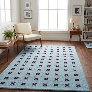 May include: A light blue area rug with a repeating pattern of dark red cross shapes. The rug is rectangular and placed on a hardwood floor. The room also features a wooden chair, side table, and a bookshelf.