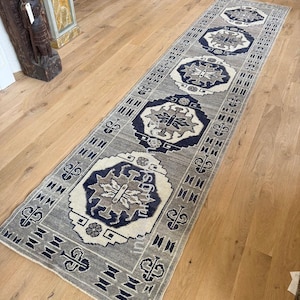 Peut inclure: Un long tapis rectangulaire gris avec un motif géométrique répétitif en bleu marine, beige et blanc. Le tapis est posé sur un plancher en bois clair, mettant en valeur son design et sa texture. Le design du tapis présente un motif de médaillon central.