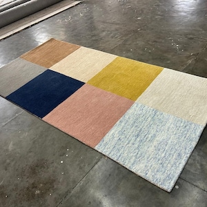 May include: Rectangular area rug with a patchwork design. The rug features squares of various colors, including tan, cream, navy blue, pink, yellow, and light blue. The rug is made of a soft material and is suitable for a living room or bedroom.