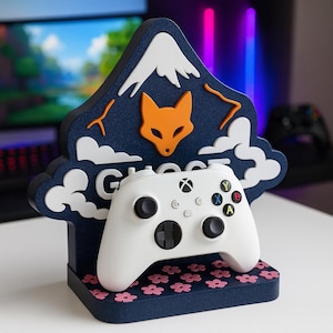 Könnte beinhalten: Ein weißer Xbox-Controller liegt auf einem marineblauen Ständer mit Berg- und Fuchsmotiv. Der Ständer hat weiße Wolken und eine rosa Blumenbasis. Das Wort "Ghost" ist sichtbar.
