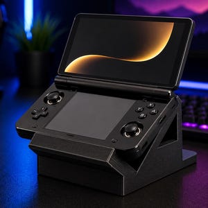 Könnte beinhalten: Schwarze Handheld-Spielkonsole mit klappbarem Bildschirm, auf einem schwarzen Ständer. Der Bildschirm zeigt ein goldgelbes, geschwungenes Design. Die Konsole hat Steuertasten und Joysticks. Das Setup befindet sich auf einer dunklen Oberfläche mit Tastatur und Kopfhörern im Hintergrund.